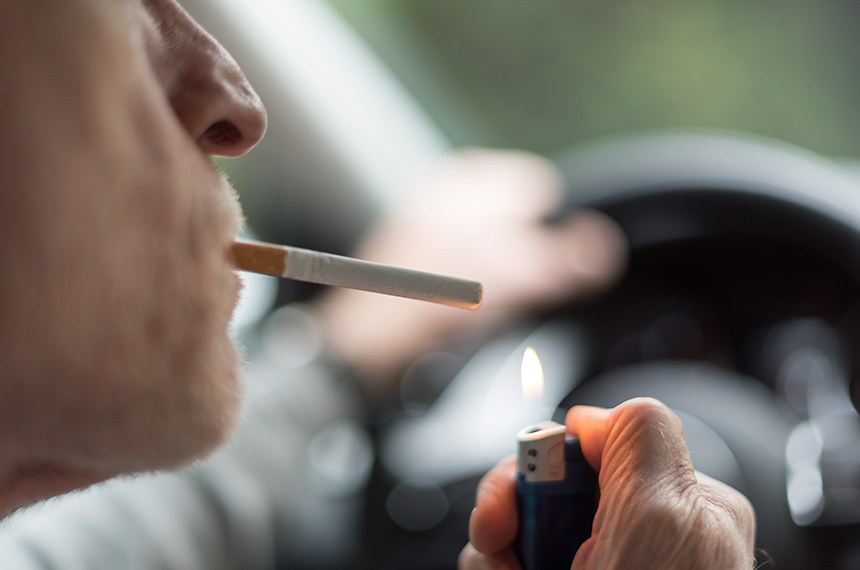 Imagem de um homem  com um cigarro na boca enquanto dirige carro.  