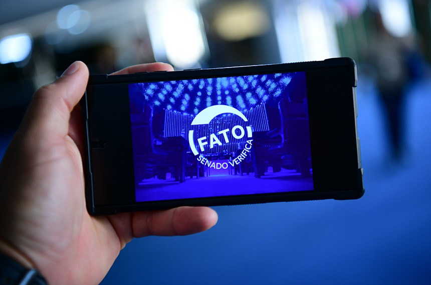 Em foco: Uma mão segurando um celular em que a tela mostra um carimbo de Fato.