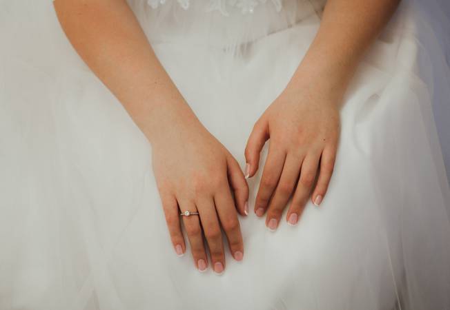 Elegant french manicure for wedding day. Bride is holding her hands with nice nails and engagement ring on white tulle puffy dress.