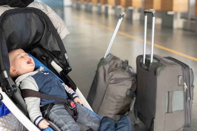 Screaming baby boy sitting in stroller near luggage at airport terminal. Child in carriage near check-in desk counter. Children tears, panic and hysterics. Travelling with small children concept.