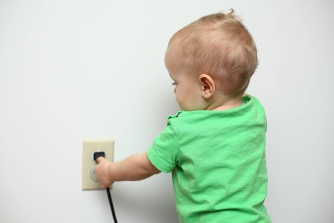 A close-up image of a Caucasian baby boy standing near the electrical outlet and playing with a power cord plugged into it. Baby trying to unplug the power cord from the wall outlet. Baby proofing, Danger and hazard for babies. Childproofing a home.