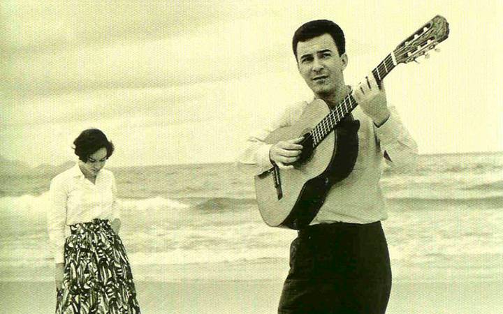 João Gilberto, em preto e branco, tocando violão na praia.