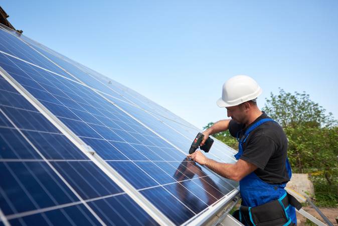 Technician connecting solar panel to metal platform using electrical screwdriver on blue sky copy space background. Stand-alone solar system installation, efficiency and professionalism concept.