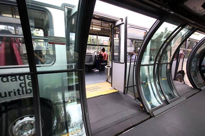 Ônibus urbano de Curitiba parado, com a porta aberta e ligada a uma estação tubo. No fundo da imagem, uma pessoa está sentada em um dos assentos do coletivo. 