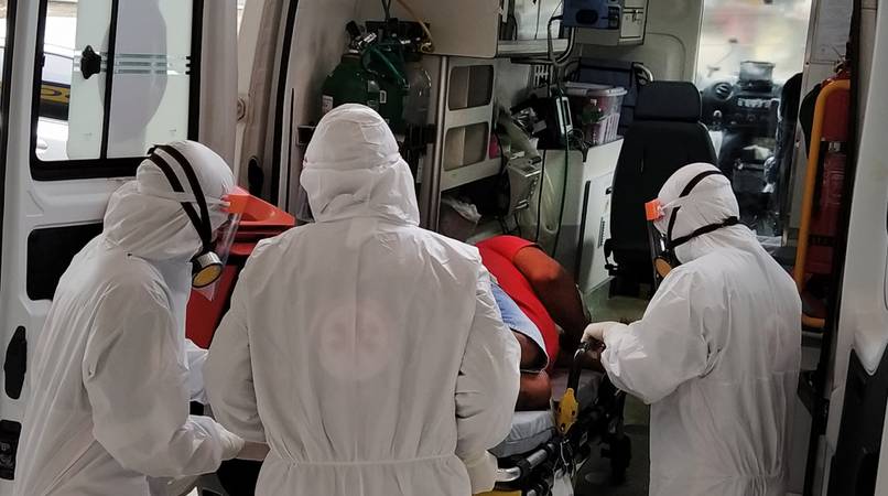 Profissionais de saúde coloca paciente, com covid-19, em ambulância. Os profissionais vestem trajes brancos de proteção, com óculos e máscaras.