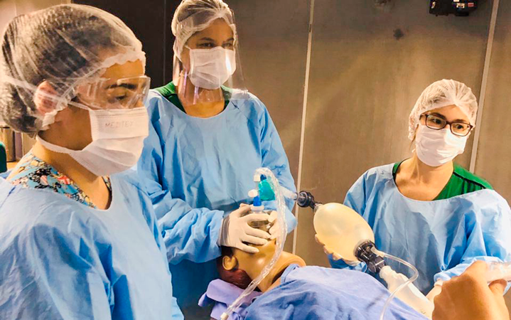 Três estudantes de medicina são instruídos sobre procedimento respiratório. Umas das estudantes segura uma espécie de respirador manual, que está sendo colocado num boneco. 