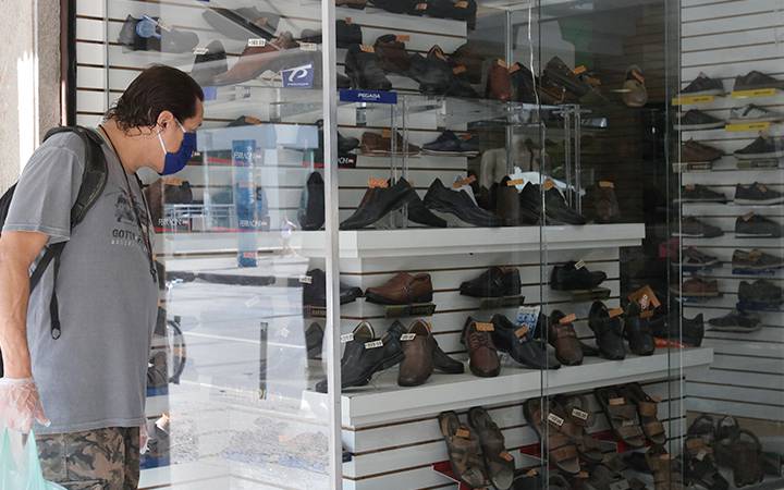 Homem, com máscara no rosto e luvas nas mãos, observa vitrine de loja de sapatos. A loja parece vazia e os sapatos são quase todos  cinza, pretos ou marrom.