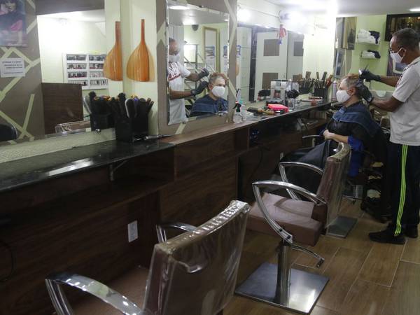 Barbearia com duas cadeiras vazias e ao fundo um profissional corta o cabelo do cliente. Ambos usam máscaras. 