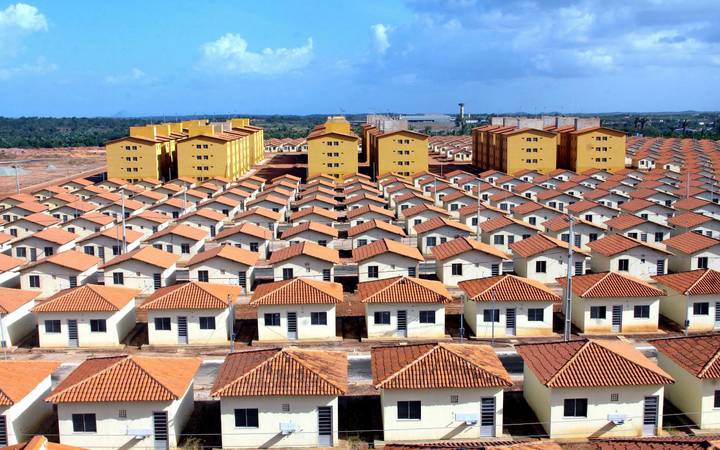 Dezenas de casas do programa habitacional Minha Casa, Minha Vida. Mais perto do horizonte, a imagem mostra o céu azul e prédios amarelos.