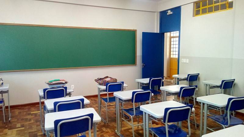Sala de aula vazia. Cadeiras azuis e mesas brancas contrastam com o piso marrom e paredes brancas. 