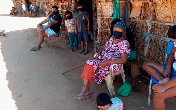 Indígenas do povo Xavantes sentados na porta da oca, usando máscaras como proteção contra a covid-19. 
