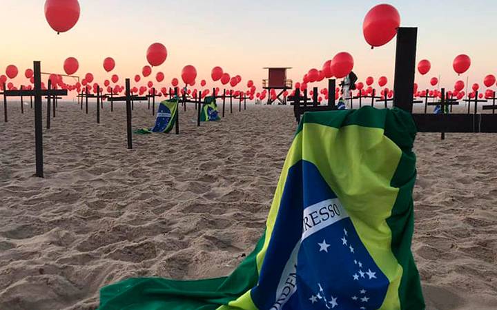 Fileira de cruzes com balões vermelhos colocados na praia para lembrar os 100 mil mortos por covid. No primeiro plano, a primeira cruz tem uma bandeira do Brasil.